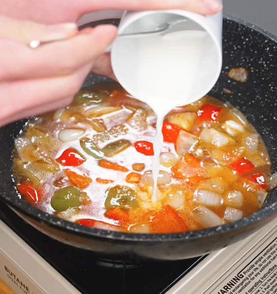 Cornstarch slurry being poured into the bubbling sweet and sour sauce as it thickens to a glossy consistency.
