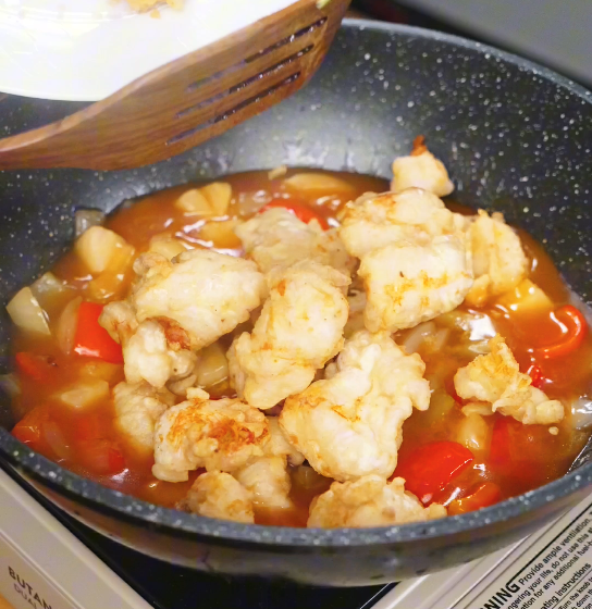 Fried chicken pieces being added back into the pan and tossed through the thick sweet and sour sauce with pineapple and vegetables until evenly coated.