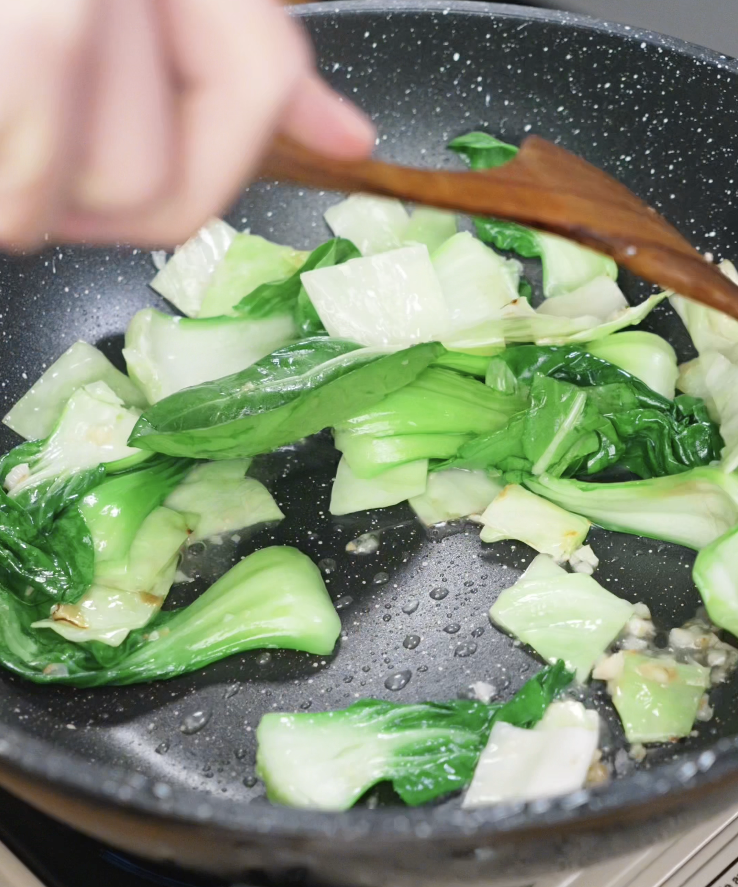Fresh bok choy leaves cooking down in a sizzling wok alongside garlic and cabbage.