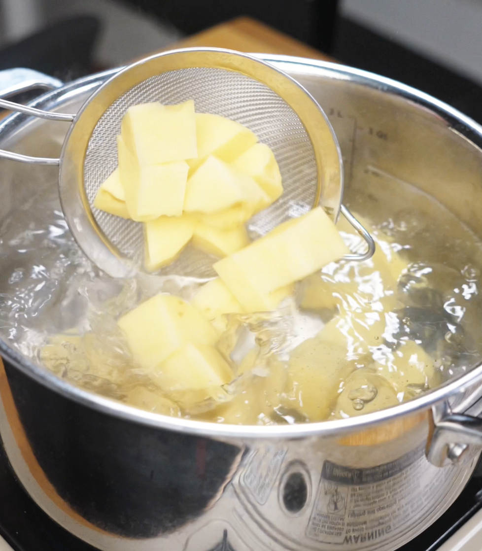 Boiling diced potatoes in a large stainless steel pot with a mesh strainer, starting the cooking process for roast potatoes.