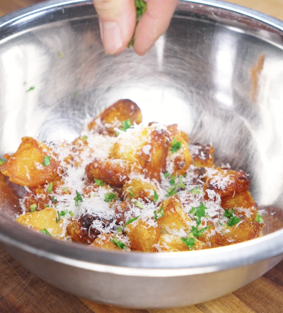Tossing crispy roasted potatoes with freshly grated parmesan cheese and chopped herbs in a metal bowl.