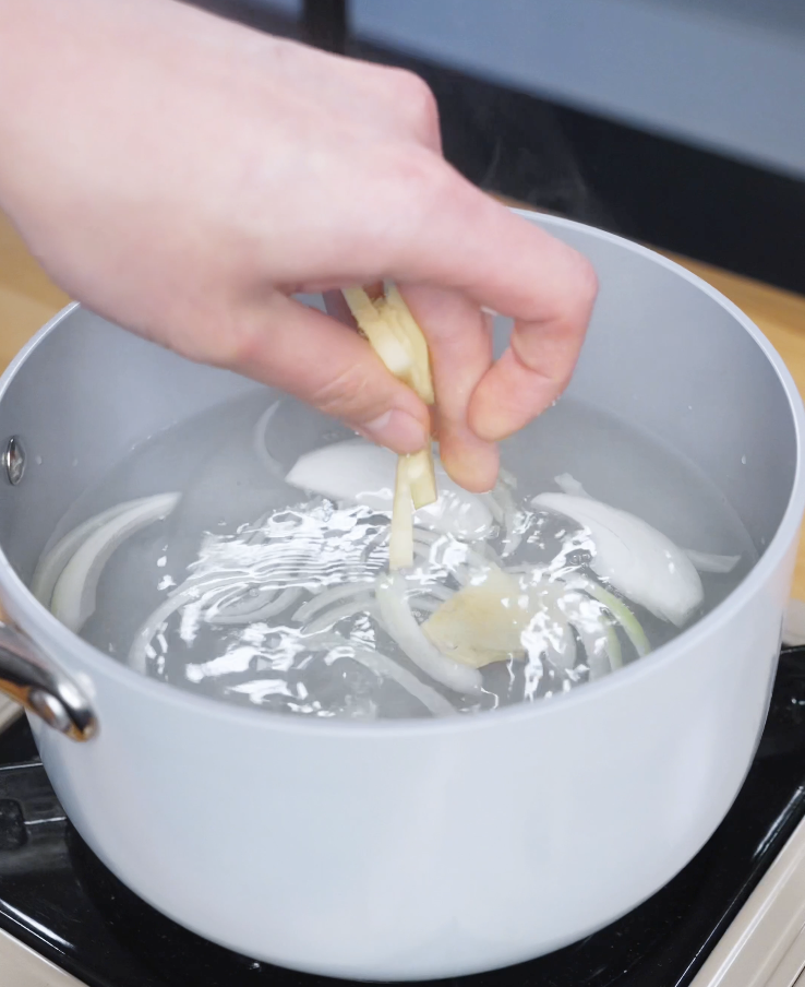 Sliced onion, ginger, and whole garlic cloves being added to a lightly salted pot of water to create a fragrant poaching liquid for juicy chicken thighs.