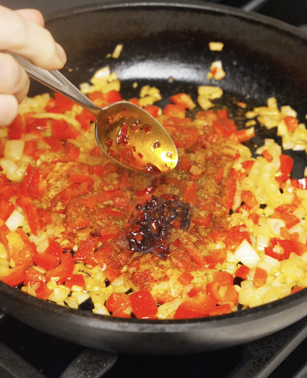 Chili oil and spices being spooned into saut&eacute;ed peppers and onions, blooming the aromatics for the shakshuka sauce.