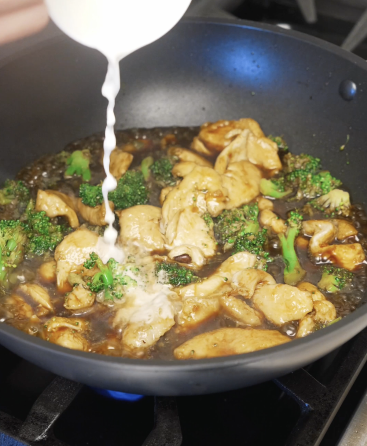 Cornstarch slurry being poured into a pan of chicken and broccoli simmering in a savory soy-based sauce, thickening it into a glossy stir fry coating.