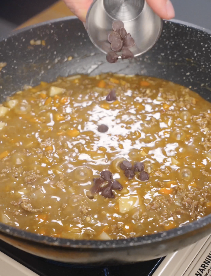 Dark chocolate being added to simmering Japanese ground beef curry to deepen flavor and add richness to the sauce.