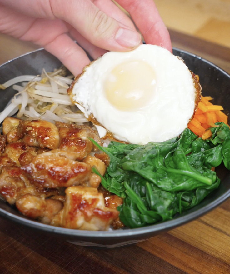Fried egg being placed on top of a chicken bibimbap bowl with chicken, vegetables, and rice before adding sauce.