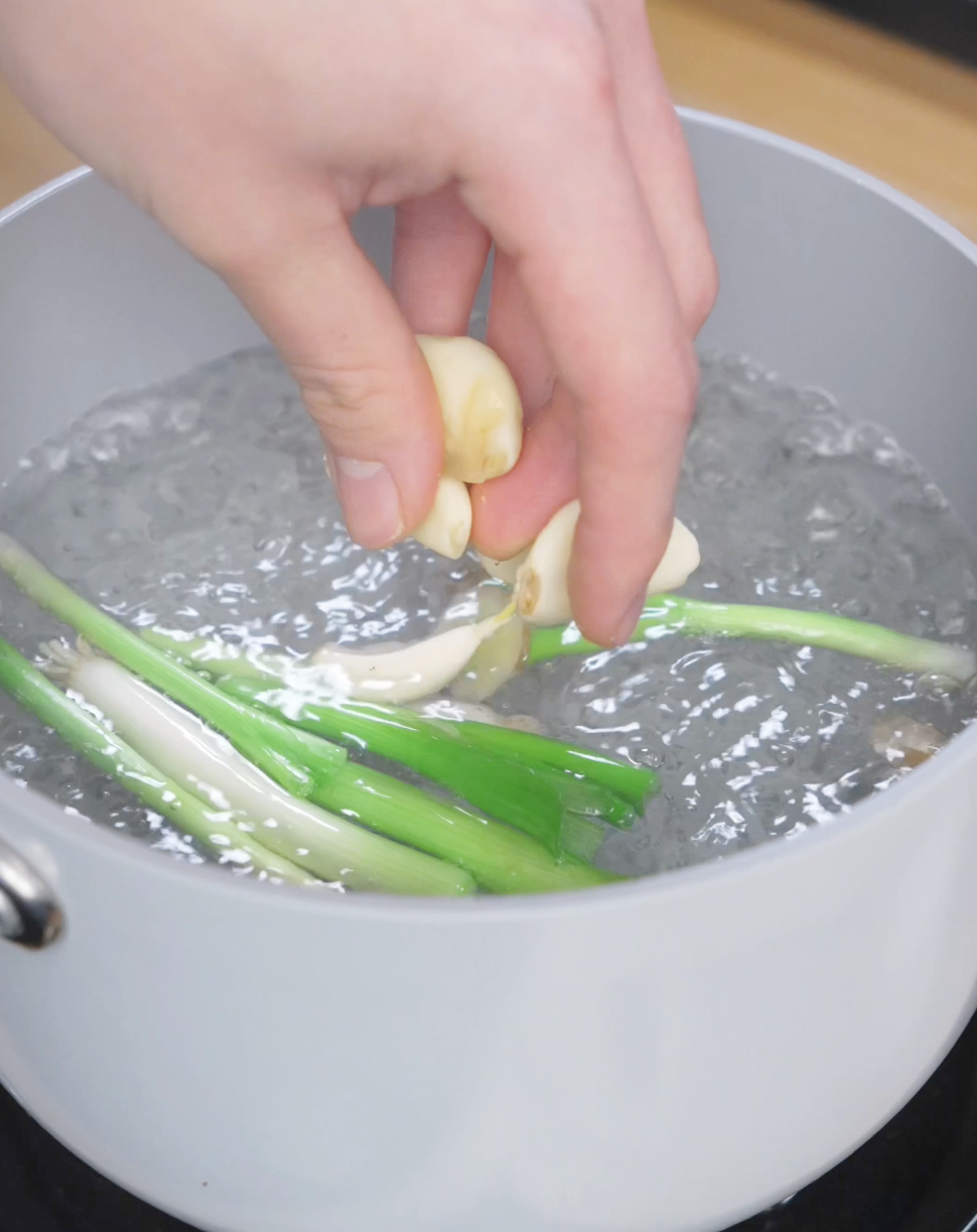 Whole garlic cloves and scallions being added to boiling water to infuse aromatics while poaching chicken for ginger scallion chicken.