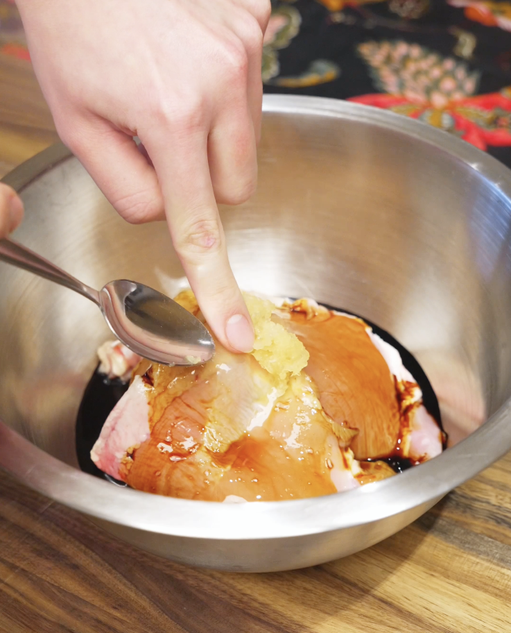 Minced lemongrass, garlic, soy sauce, fish sauce, and sugar being added to raw chicken thighs in a metal mixing bowl for lemongrass chicken marinade.