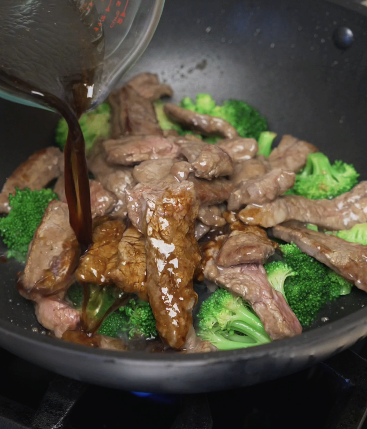 Savory beef and broccoli sauce being poured over cooked beef and broccoli in a pan, starting the final simmer.