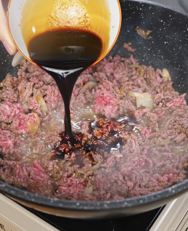 Dark soy-based Pad Kra Pao sauce being poured into browned ground beef, creating a glossy, umami-rich coating.