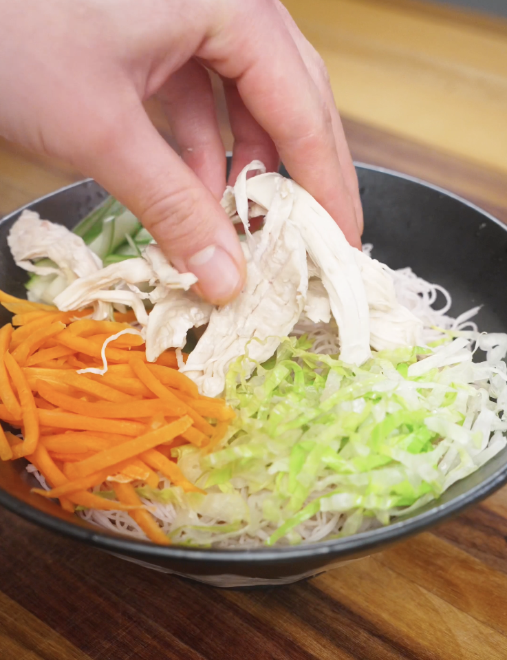 Shredded poached chicken being placed on top of rice noodles, lettuce, carrots, and cucumbers in a Vietnamese chicken salad bowl.