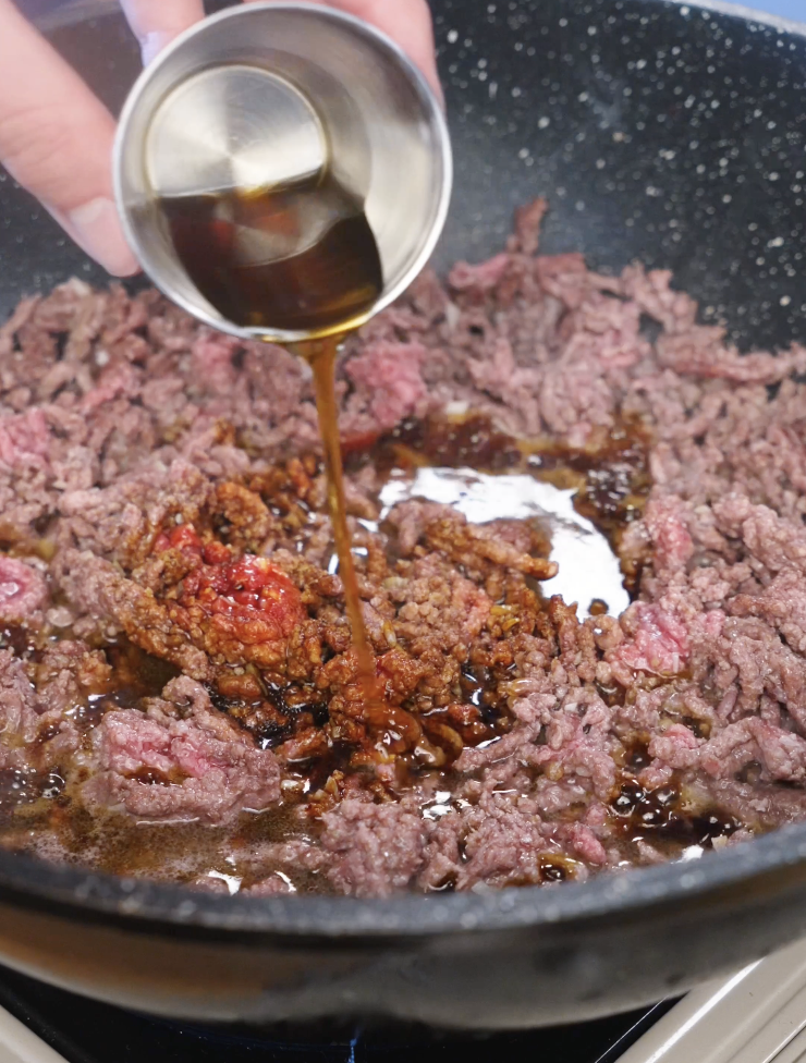 Dark and light soy sauce being poured into browned ground beef to create the savory-sweet Korean BBQ beef sauce.