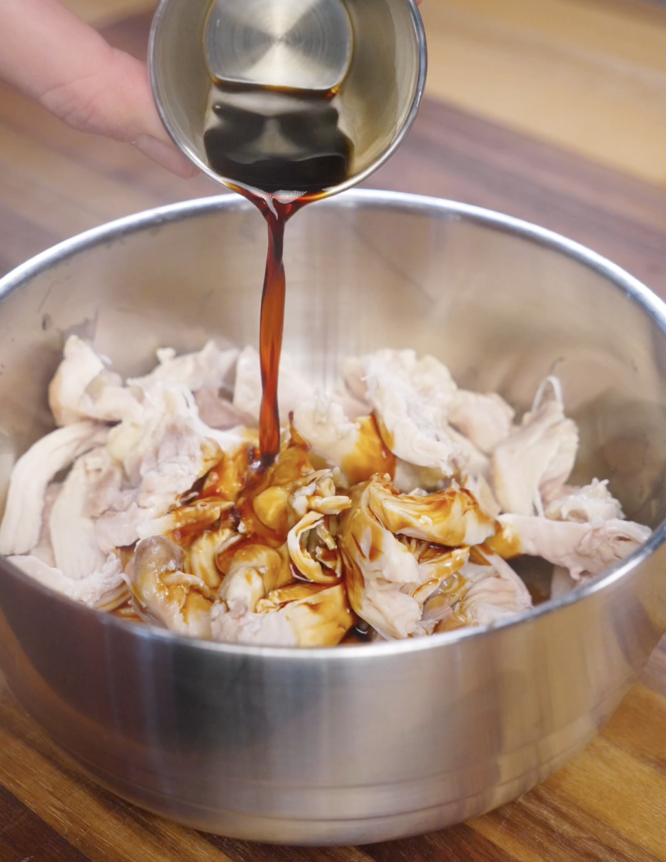 Dark soy sauce being poured over shredded poached chicken in a mixing bowl to season ginger scallion chicken before serving.