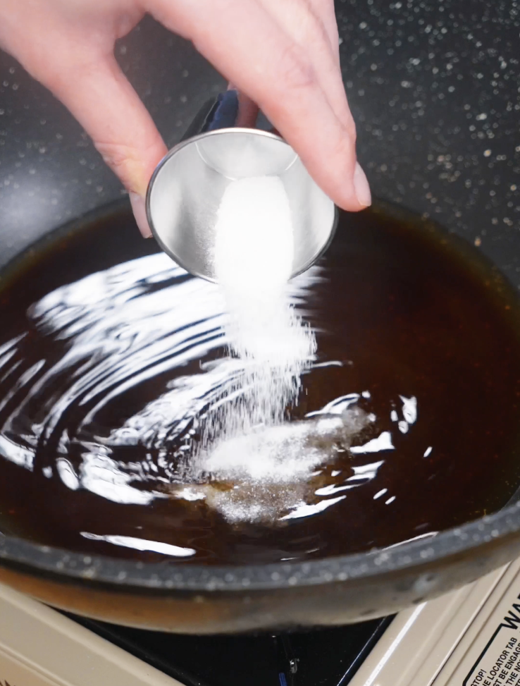 Pouring sugar into a pan of simmering soy-based gyudon broth to balance the savory and sweet flavors of the Japanese beef bowl.