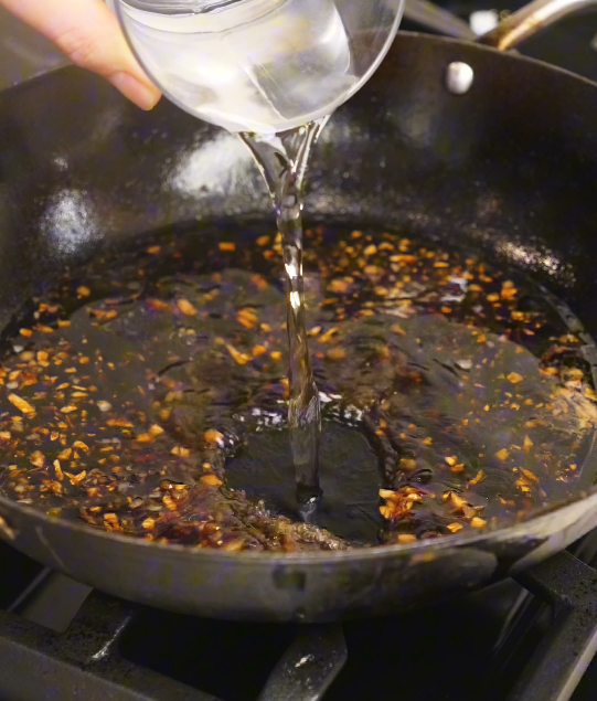 Water being poured into a pan with soy sauce, garlic, ginger, and sugar, forming the base of a homemade teriyaki sauce.