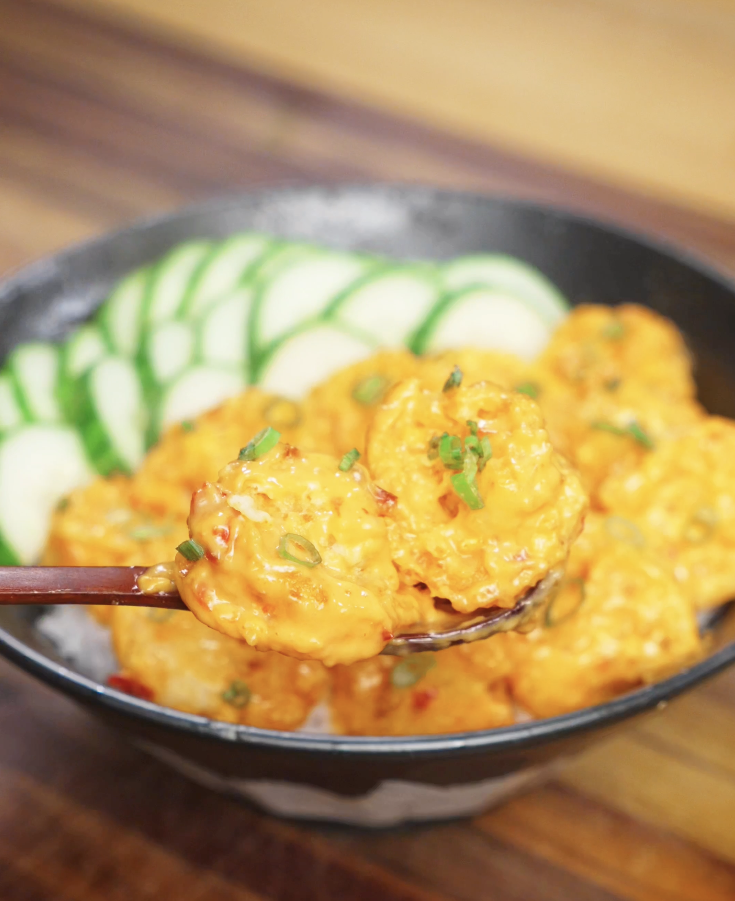 Close-up spoonful of crispy Bang Bang Shrimp coated in creamy sweet-spicy sauce, lifted from a rice bowl with cucumbers in the background.