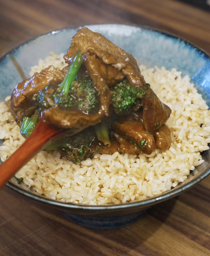 Saucy beef and broccoli being spooned over a bowl of steamed rice, finishing the classic beef and broccoli dish.