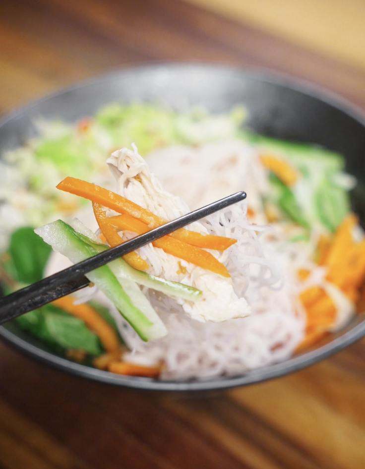 Chopsticks lifting shredded poached chicken with rice noodles, julienned carrots, and cucumbers from a Vietnamese chicken salad bowl.