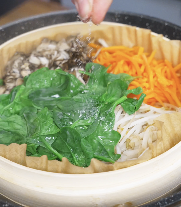 Assorted vegetables including spinach, bean sprouts, carrots, and mushrooms arranged in a steamer basket as the prep step for a healthy chicken bibimbap bowl.