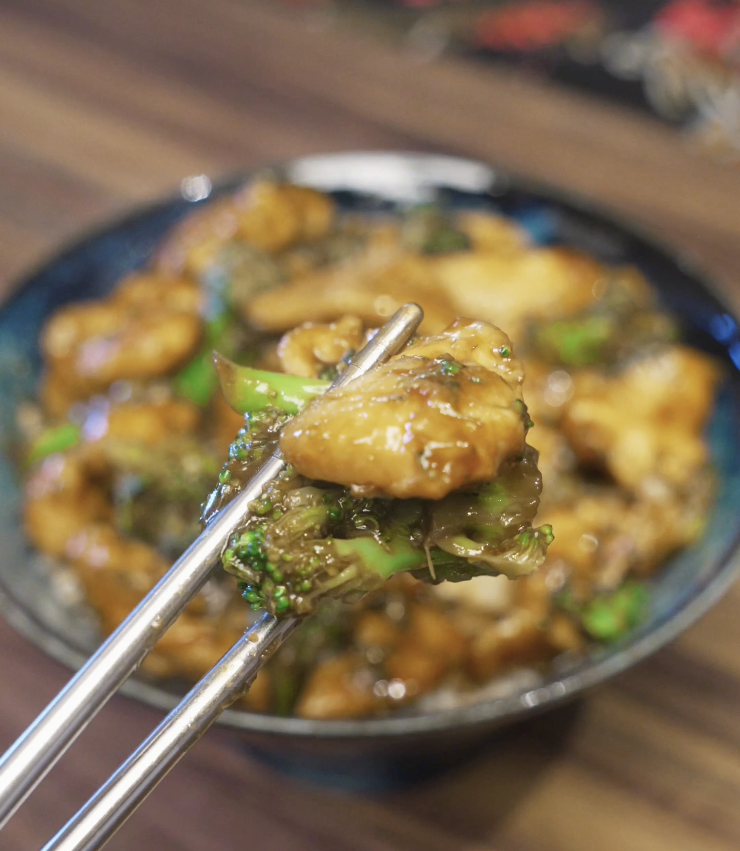A glossy bite of chicken and broccoli held with chopsticks, showing tender chicken coated in a savory soy-based sauce and bright green broccoli florets from the finished chicken and broccoli dish.