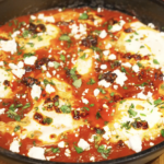 Chili oil shakshuka simmering in a skillet with gently poached eggs set in a rich tomato sauce, finished with feta, herbs, and chili oil.