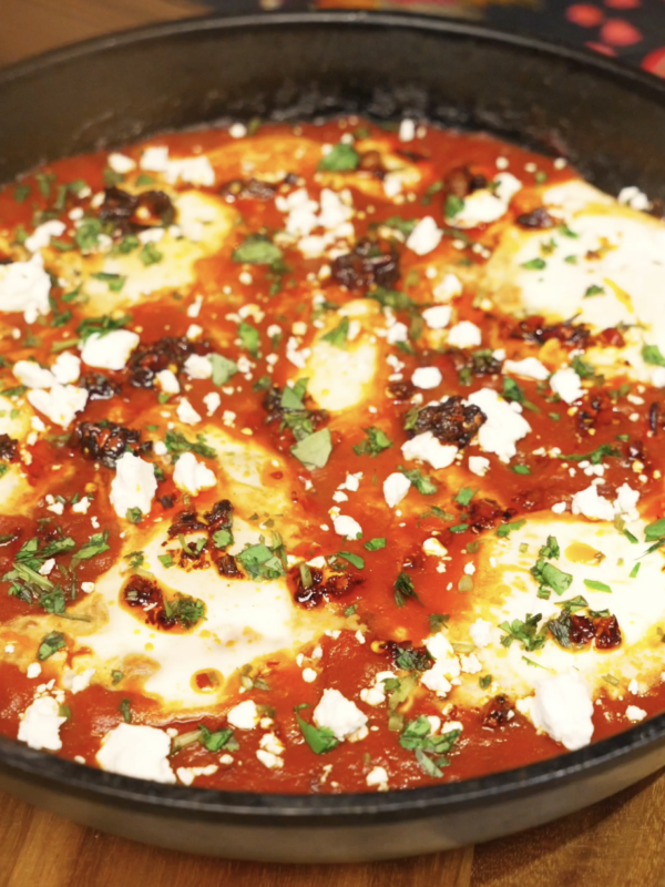 Chili oil shakshuka simmering in a skillet with gently poached eggs set in a rich tomato sauce, finished with feta, herbs, and chili oil.