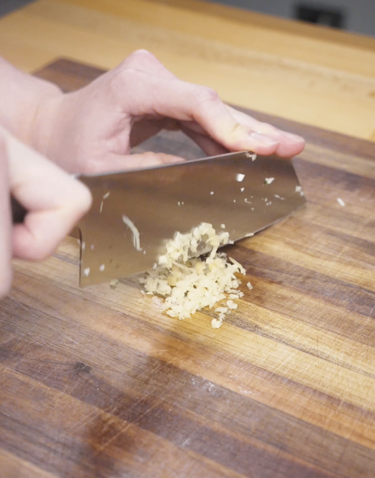 Fresh garlic being finely chopped on a wooden cutting board to build the aromatic base for Spicy Peanut Chicken.