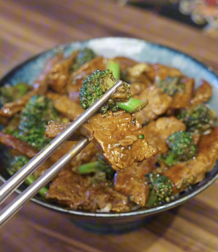 Chopsticks holding a piece of tender beef and broccoli coated in savory sauce above a bowl of beef and broccoli.