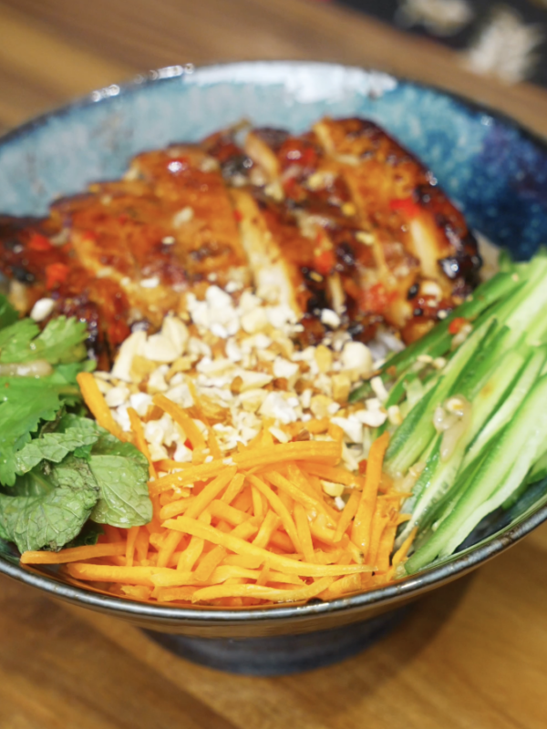 Chopsticks lifting a bite of lemongrass chicken with vermicelli noodles, herbs, and vegetables from a Vietnamese-style chicken bowl.