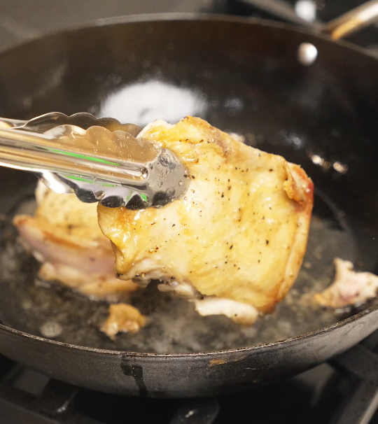 Chicken thighs being flipped in the pan with tongs, revealing golden, crispy skin after searing over medium heat.