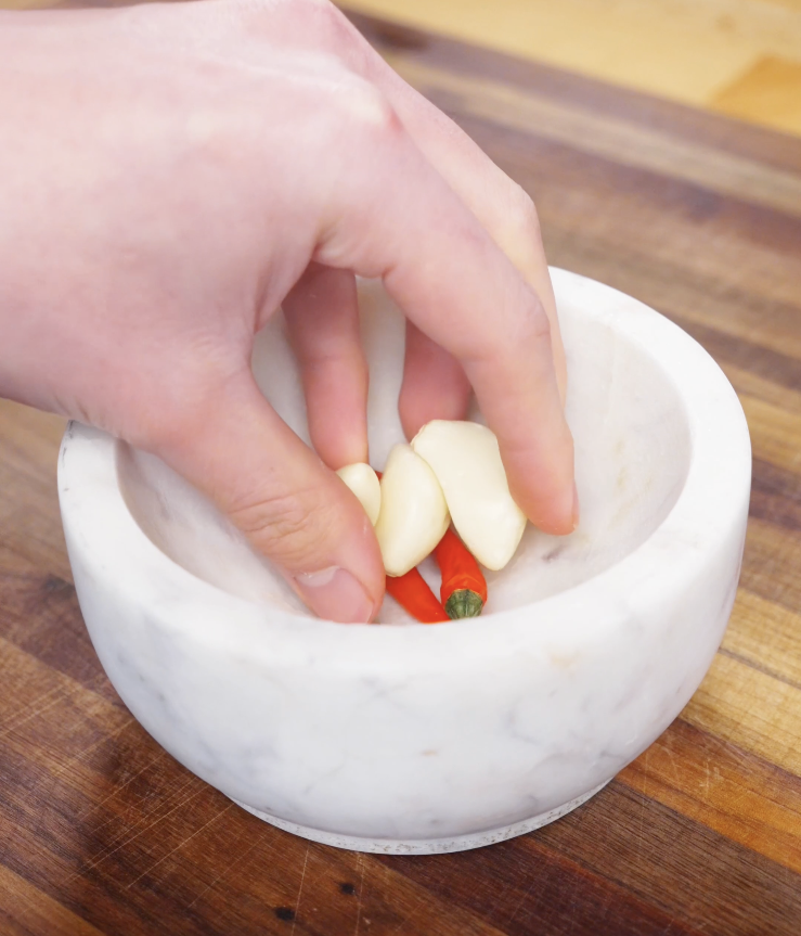 Garlic cloves and Thai chilies being crushed in a mortar and pestle to release their aroma for authentic Pad Kra Pao flavor.