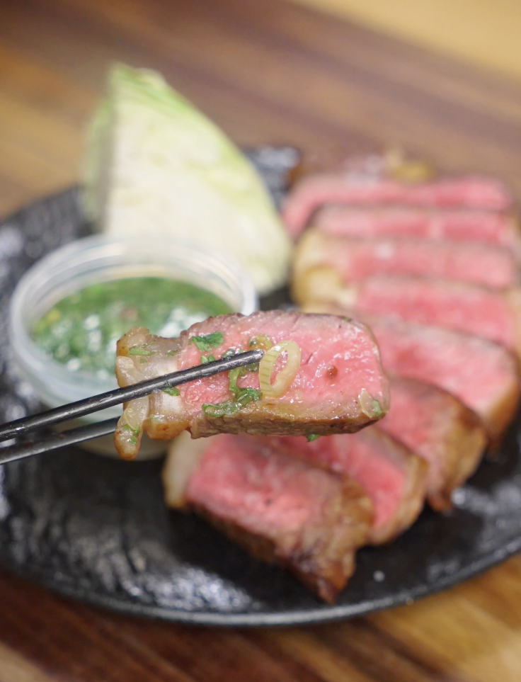 Close-up of a chopsticks bite of medium-rare Crying Tiger Steak dipped in Thai-style sauce, showing juicy steak and fresh herbs.