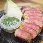 Finished Crying Tiger Steak sliced medium-rare and served with a Thai-style dipping sauce, fresh cabbage, and flaky salt on a black plate.