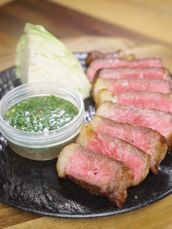 Finished Crying Tiger Steak sliced medium-rare and served with a Thai-style dipping sauce, fresh cabbage, and flaky salt on a black plate.