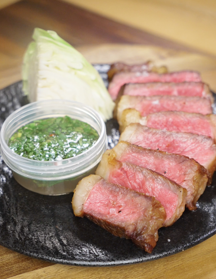 Finished Crying Tiger Steak sliced medium-rare and served with a Thai-style dipping sauce, fresh cabbage, and flaky salt on a black plate.
