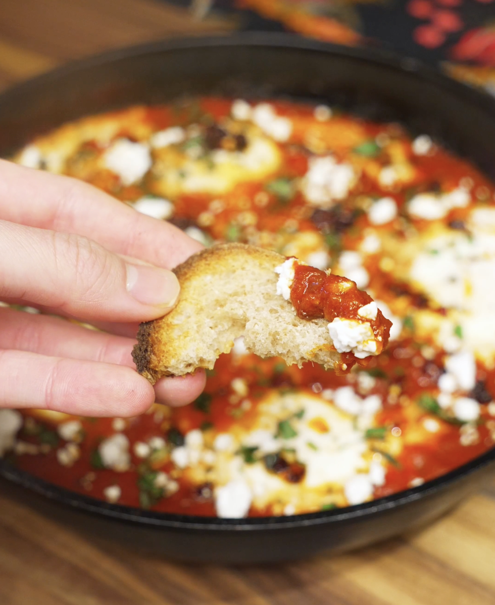 A piece of toasted bread dipped into chili oil shakshuka, scooping up tomato sauce, soft egg, and crumbled feta for a perfect bite.