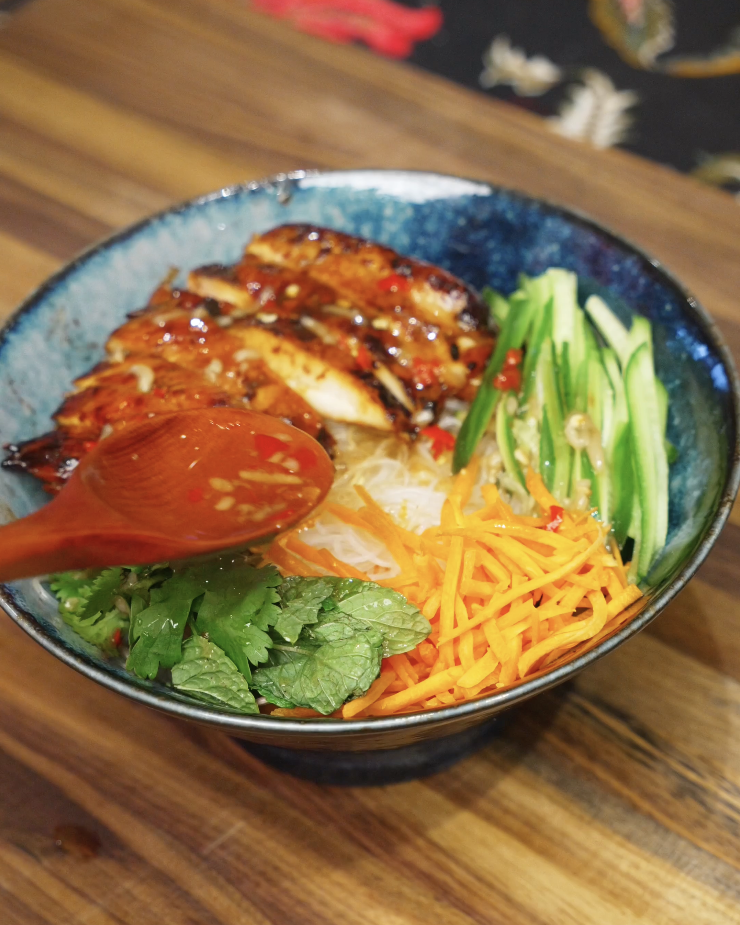 Spoon drizzling tangy nuoc cham sauce over a lemongrass chicken vermicelli bowl with fresh herbs and vegetables.