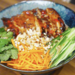 Vermicelli noodles topped with sliced lemongrass chicken, cucumber, shredded carrots, mint, and cilantro in a ceramic bowl.