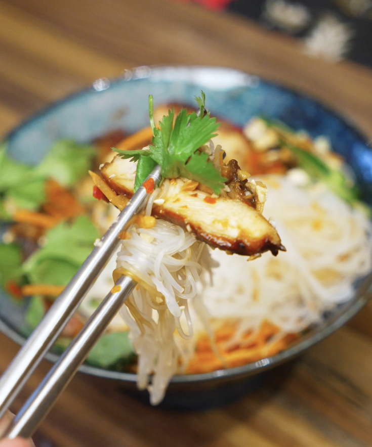 Chopsticks lifting a bite of lemongrass chicken with vermicelli noodles, herbs, and vegetables from a Vietnamese-style chicken bowl.