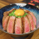 Thinly sliced medium-rare steak arranged over rice and topped with scallions and a raw egg yolk in a ceramic bowl.