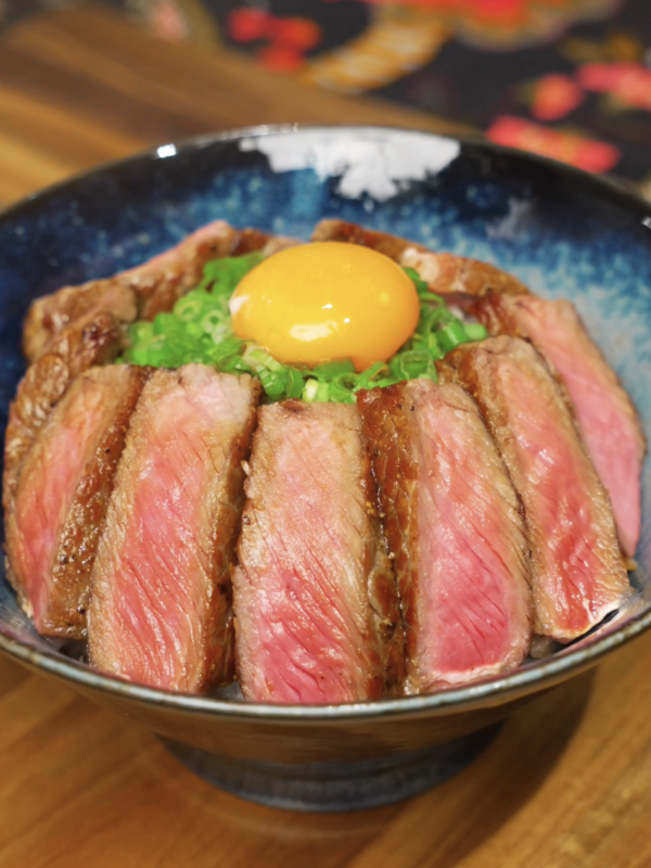 Thinly sliced medium-rare steak arranged over rice and topped with scallions and a raw egg yolk in a ceramic bowl.