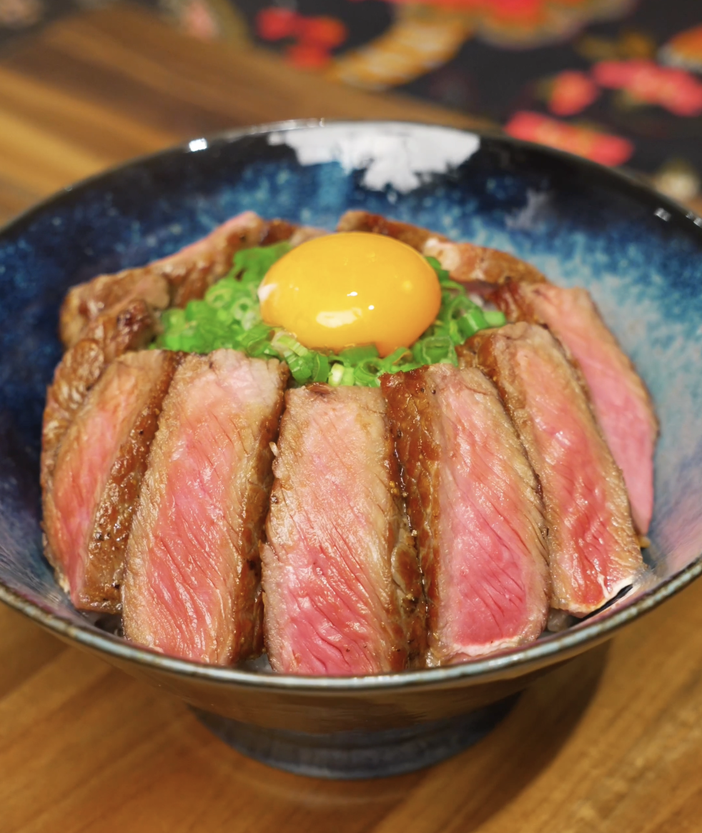 Thinly sliced medium-rare steak arranged over rice and topped with scallions and a raw egg yolk in a ceramic bowl.