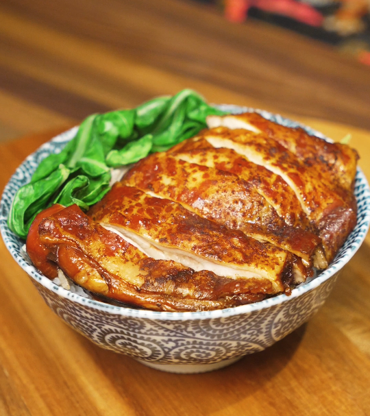 Soy sauce chicken sliced and served over steamed white rice with glossy sauce and leafy greens in a patterned bowl.