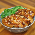 Teriyaki chicken sliced and served over steamed rice with glossy sauce, sesame seeds, and steamed greens in a patterned bowl.