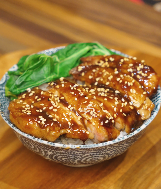 Teriyaki chicken sliced and served over steamed rice with glossy sauce, sesame seeds, and steamed greens in a patterned bowl.