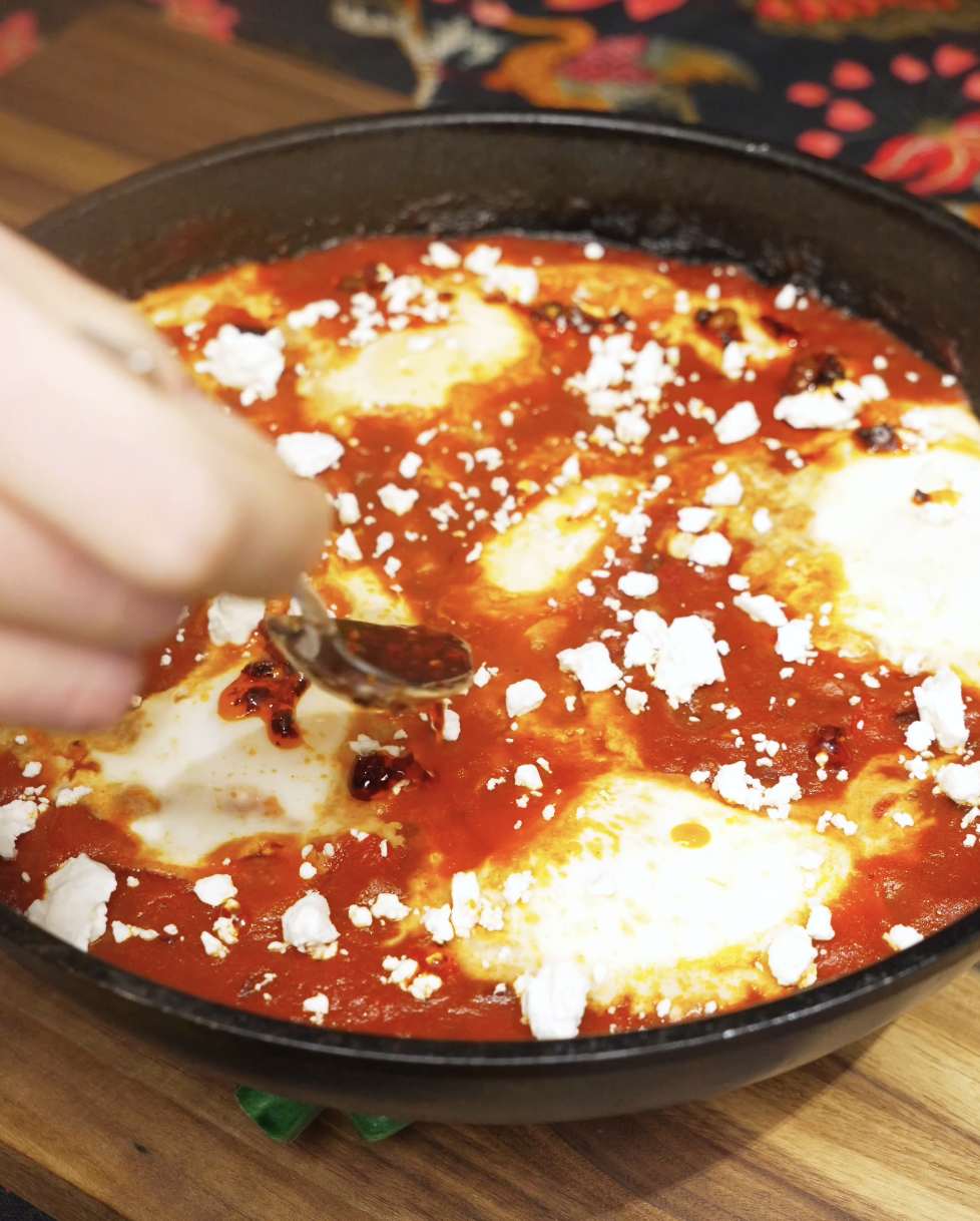 Chili oil being drizzled over shakshuka with poached eggs, crumbled feta, and herbs as the final garnish before serving.