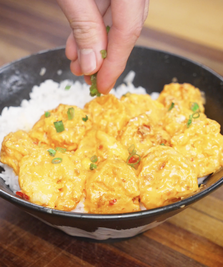 Freshly chopped scallions being sprinkled over Bang Bang Shrimp coated in creamy sauce and served over rice.