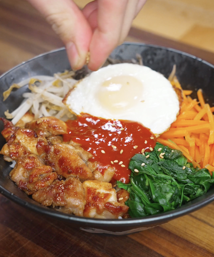 Sprinkling sesame seeds over chicken bibimbap with gochujang sauce, fried egg, and vegetables as the final garnish step.