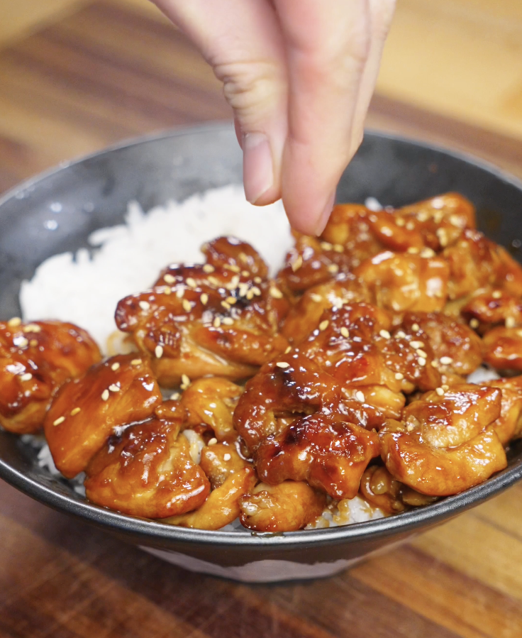 Hand sprinkling sesame seeds over freshly cooked honey garlic chicken resting on steamed rice, highlighting the sticky glaze and final plating step.