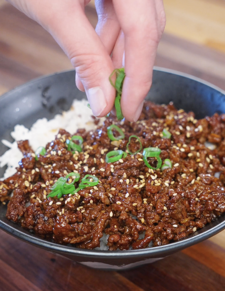 Fresh sliced scallions being sprinkled over Korean BBQ beef and rice to finish the dish with color and freshness.